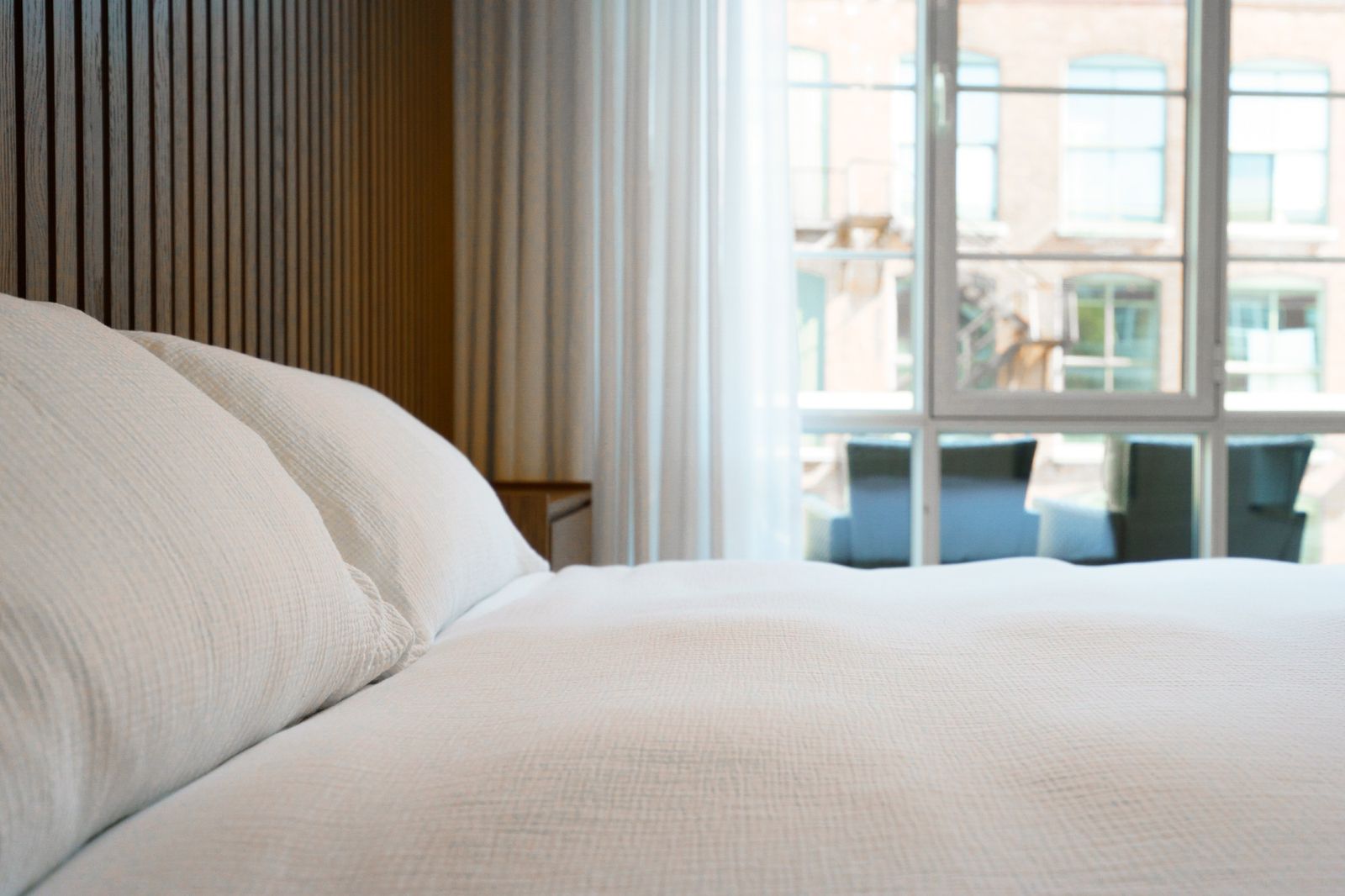 A bed with white linen by a large window, a brick building across the street